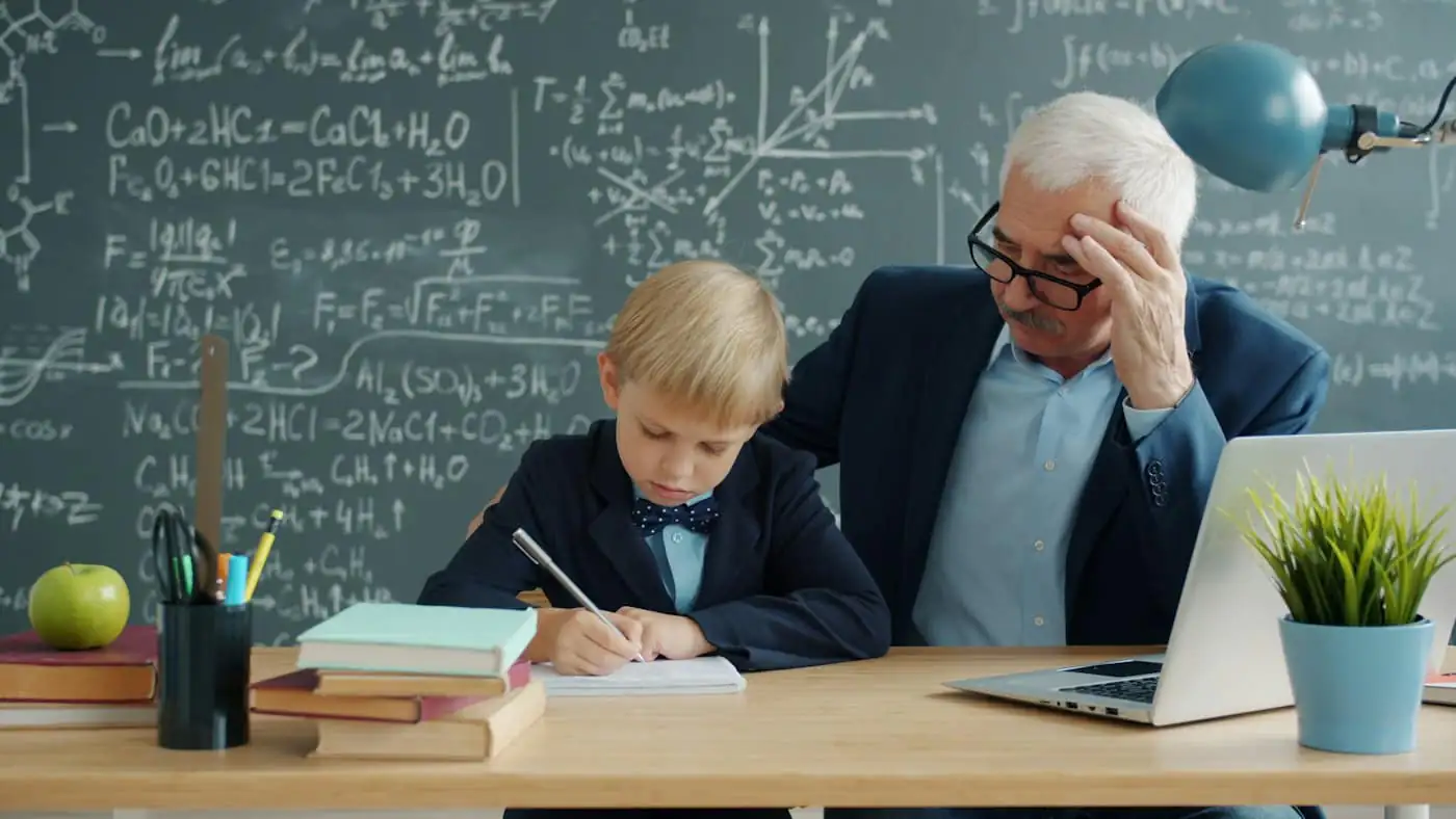A teacher sitting with a young student working at a desk in front of a chalkboard
